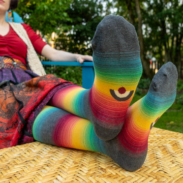 A woman wearing vibrant rainbow-striped knee socks, seated outdoors with her feet up to reveal the ModSocks knit-in logo on the sole.