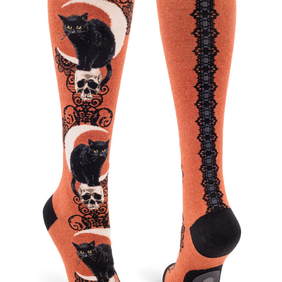 Rust orange knee-high socks with black cats, skulls, crescent moons and lace patterns as well as a faux lace ribbon back seam.
