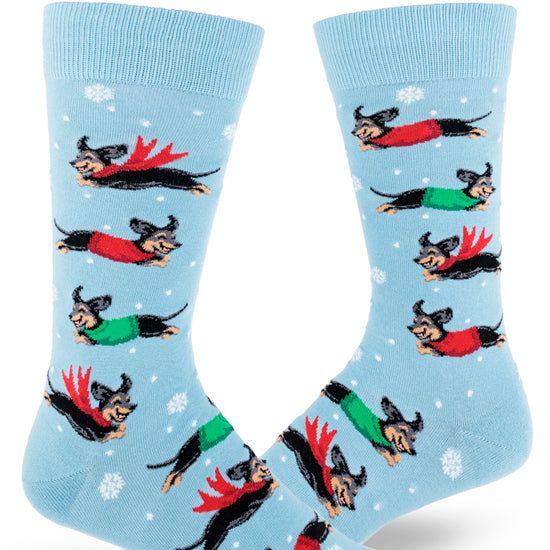 Light blue crew socks for men with black and tan dachshunds wearing red and green scarves and sweaters and jumping between snowflakes.
