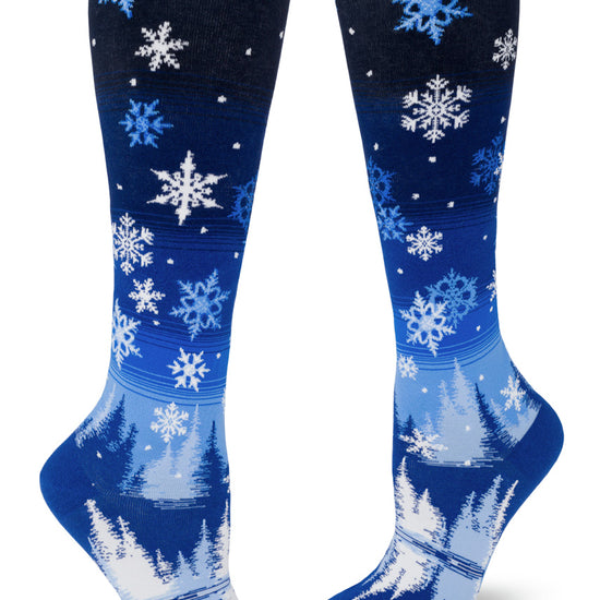 Blue knee-high socks with an allover pattern of falling snowflakes above a scene of a peaceful forest and lake in nighttime.