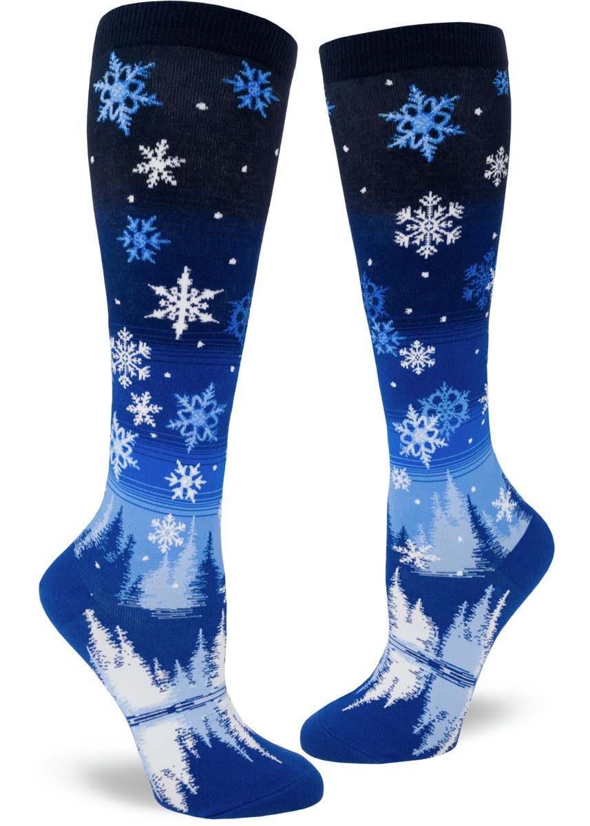 Blue knee-high socks with an allover pattern of falling snowflakes above a scene of a peaceful forest and lake in nighttime.